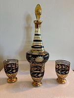 Full view of the etched glass decanter with stopper and three matching glasses surrounding it on a flat white surface against a white wall.
