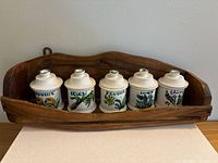 Front view of five ceramic jars with lids placed on wooden shelf, each jar hand-painted and labeled in French with herbs and spices