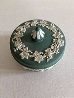 Top view of green Wedgwood jasperware trinket dish lid showing white floral and leaf relief decoration in circular pattern and central knob with white accents.