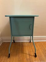 Front view of aqua metal nightstand with closed cabinet door and four legs on castors.