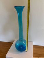 Tall blue blown glass vase measured against a tape measure, approximately 18 inches tall, standing on a wooden surface against a beige wall.