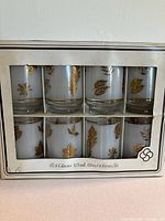 Front view of the set of 8 drinking glasses in original packaging. Glasses have frosted clear glass bodies with gold leaf motif and gold trim accents.