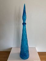 Front view of tall blue glass genie bottle with wave texture and pointed stopper.