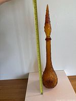 Full height photo showing tall amber glass bottle with slender neck, textured body, and pointed stopper with wave pattern, alongside measuring tape indicating approximately 20 inches tall.