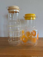 Two vintage Pyrex juice containers side by side: one clear with measurement markings, one decorated with orange slice graphics and yellow lid.