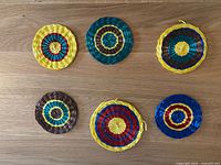 Six brightly colored round raffia drink coasters arranged on wooden surface, showing various concentric circle color patterns