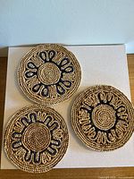 Three round woven raffia trivets laid flat, showing natural and navy color spiral braid designs with looped accents.