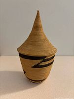 Front view of the small handwoven conical basket with a pointed lid, showing tan weave with black patterning.