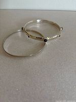 Two sterling silver bangles shown side by side on flat surface, one hammered with black/brown stones and one smooth and thin.