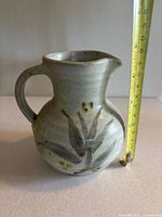 Front view of pottery vessel showing rounded body, handle, spout, and brown leaf-like decorative brush strokes on off-white matte speckled glaze.