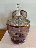 Overall view of the amethyst colored glass egg decanter set with shot glasses inside, showing hand-painted floral decorations and gilded accents