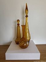 Three amber glass pieces including two tall decanters with stoppers and one hobnail vase arranged on a white box.
