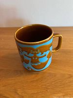 Front view of the blue mug showing the brown mermaid and fish design, with brown interior and handle.