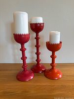 Three vintage IKEA metal candle holders in bright red and orange colors displayed with white pillar candles on a wooden surface.