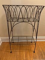 Full front view of vintage metal patio planter basket stand showing decorative curved metal arches and lower horizontal slat shelf.