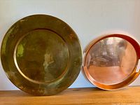 Photo of both trays side by side on the floor, large brass tray on left showing tarnish, smaller copper tray on right with reflective shine and handles visible