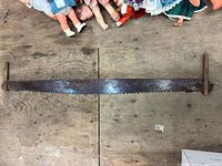 Full length view of the antique 2-man crosscut saw lying flat on a wooden floor under some dolls. Shows entire blade and handles, highlighting length and blade teeth.