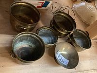 Six brass plant pots displayed on a wooden surface, showing various sizes and patina