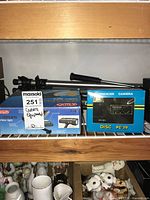 Photo showing the boxed Optex cordless video light, Premier Disc PC-20 camera box, and multiple tripods placed on a wire rack shelf.