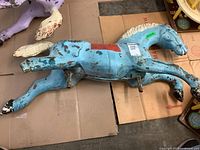 Full side view of the blue enameled metal carousel horse laying on cardboard surface showing extensive paint loss and missing hind hoof with detached tail in background.