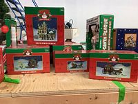 Seven boxes of Santa's Trim Shoppe Christmas porcelain lighted houses and carriages arranged on a wooden surface.