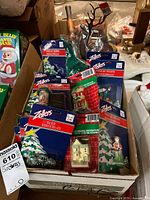 Overview of a box containing multiple Christmas village accessories, mostly in original Zellers brand packaging with blue and red color scheme, featuring a Christmas tree graphic. Items include figurines, bushes, house decorations, and some unpackaged lamp posts.