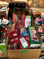 Box containing assorted Christmas-themed items including several Santa Claus banks, a plush dog with a Santa hat, and various ornaments and boxed items