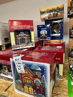 Multiple boxed hand-painted porcelain lighted Christmas village buildings stacked showing product packaging and designs