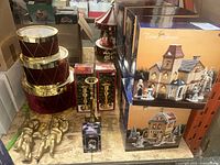 Photo of stacked red decorative drums with gold trim, gold brass candle holders in boxes, two gold cherub figurines, Carousel bell box, and two boxed Trim Shoppe Christmas village buildings.