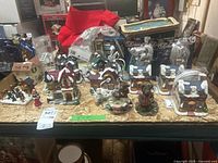 Full display of Christmas village buildings, figures, and boxed accessories arranged on wooden surface. Variety of snow-covered house models, figures, and accessories visible.