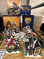 Overview photo showing five ceramic Christmas village houses with attached white electrical cords and original orange and blue Trim Shoppe boxes in the background.