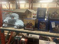 Overall view of two Holiday Mountains village display pieces on left and five Trim Shoppe Christmas village building boxes on right on a shelf.