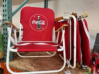 4 red Coca Cola branded folding beach chairs with white metal frames and wooden armrests stacked and displayed on a shelf, showing overall lot contents.