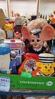 Shows a collection of colorful figural coin banks including a large figure with big ears and graduation cap, a yellow bank with striped hat, and a slot machine bank.