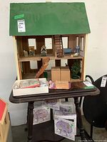 Photo of wooden doll house on table with green roof, two floors, visible staircase damage, miniature furniture and accessories on shelves and table