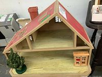 Front and side view of wooden doll house on display, showing open interior, chimney, and small tree and cabinet accessories nearby.