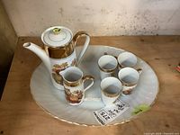 Full view of coffee pot, creamer, four espresso cups arranged on a large white scalloped oval milk glass tray with gilded edge.