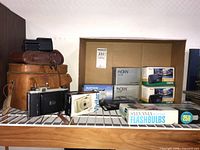 Boxed Meikai AR-4400 cameras, Wide Pic Panoramic cameras, vintage flip-front medium format camera, several leather camera cases, flash cubes and Stania flashbulbs on shelf.