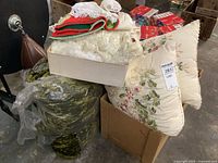 Photo showing box of lace doilies, folded lace bed linens, floral patterned pillows, and a green patterned fabric item in plastic.
