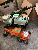 Two metal toy vehicles placed on a concrete floor: a white crane truck and an orange tractor with side attachment