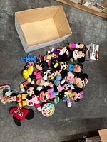 Photo showing assorted plush Disney characters including several Mickey and Minnie Mouse plushies in various colorful outfits and styles, placed on the floor next to an empty box.