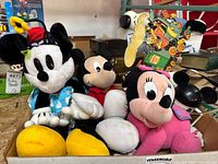 Three Disney plush dolls: Minnie Mouse in blue polka dot dress and yellow shoes, Mickey Mouse in red shirt, and Minnie Mouse in pink outfit with bow alongside a Disney cup.