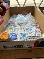 Box filled with various novelty souvenir and collectible pins, buttons, and badges wrapped in plastic.