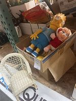Box containing Bart Simpson plush toy, Baby Miss Piggy plush, and a doll resembling Miss Piggy in red, with other toys partially visible inside the box.
