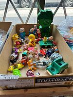 Box containing assorted small collectible character figurines including Simpsons, Muppets, California Raisins, and Peanuts, as well as some small toy vehicles and a wooden truck toy.