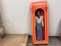 Front view of boxed Sonny Bono doll over 12 inches tall, dressed in white top and blue jeans, in an orange box with window.