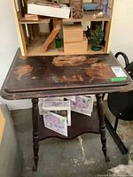 Photo of vintage wooden side table showing top surface damage and lower shelf, with nearby objects in background.