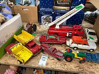 Overview of various vintage toy trucks and tractors arranged on plywood board, showing multiple toy vehicles in red, yellow, white, green, and orange with visible wear and scratches.