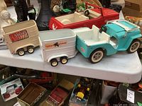 Wide view of the turquoise Jeep, red truck, and attached Safari and Tomah Stables trailers on a white surface among other items.