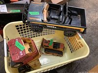 Three Tonka vehicles including orange dump truck, black and yellow pickup truck in a plastic basket on concrete floor